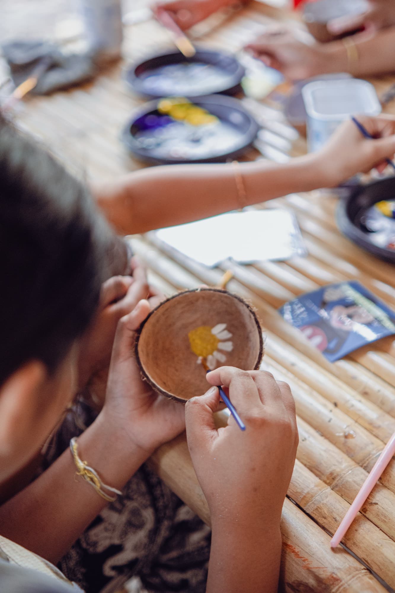 Two hands painting a yellow and white flower inside a coconut shell bowl with a fine brush.
