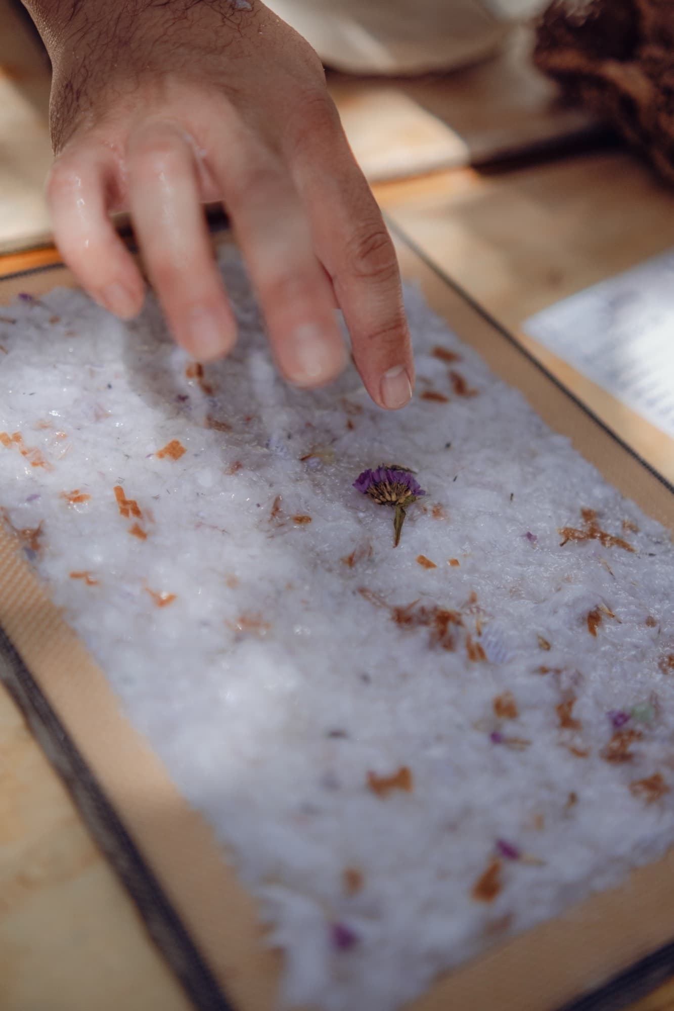 A hand presses dried petals into wet paper pulp held in a rectangular screen frame.