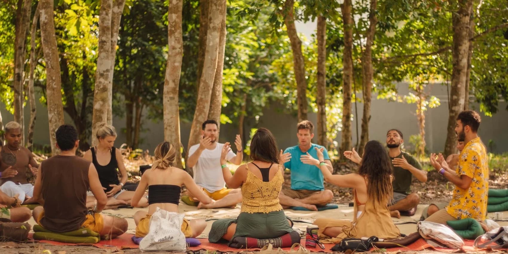 A group sits cross-legged in a circle on mats under trees while one person leads from the far side.
