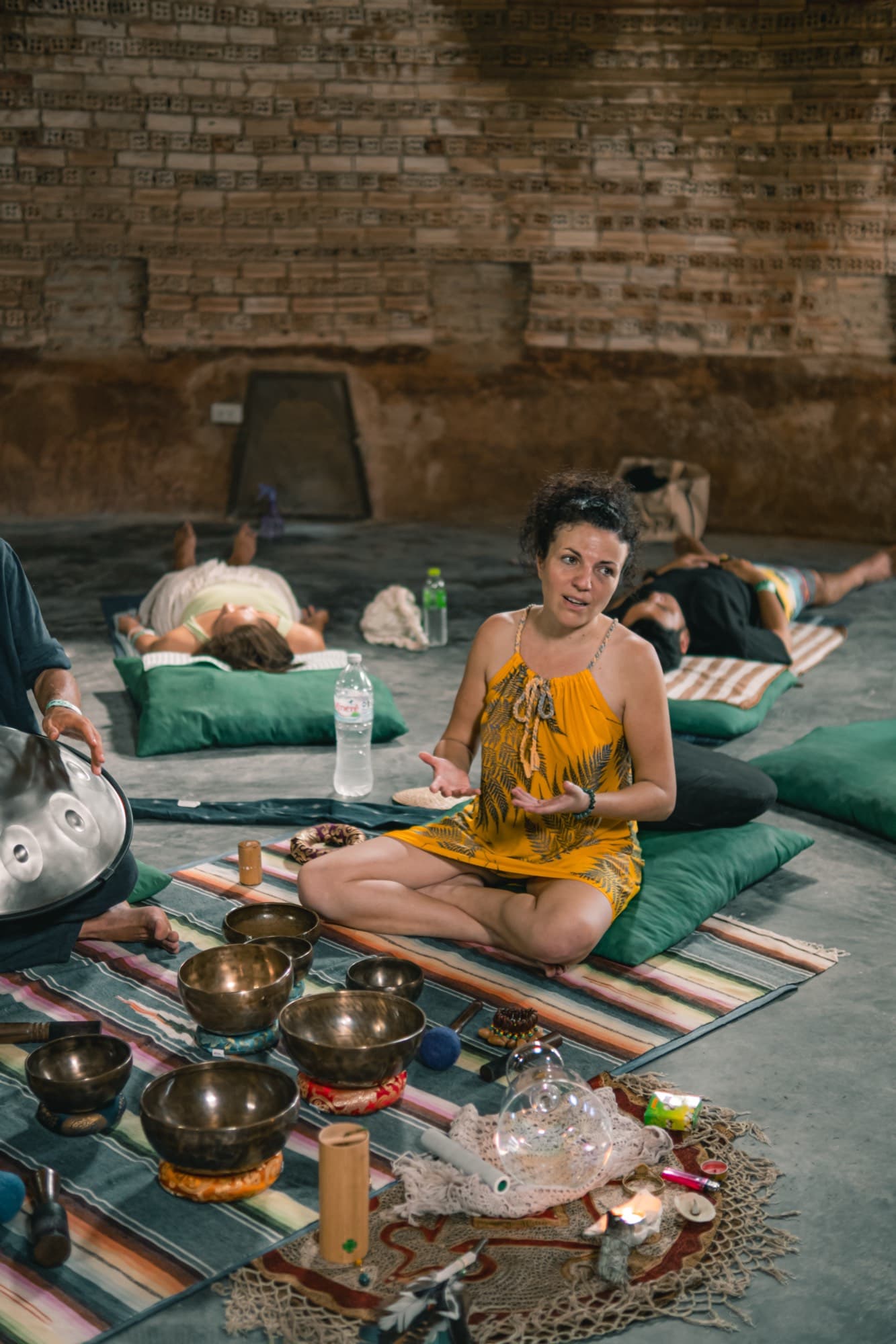 A facilitator sits cross legged beside singing bowls while people rest on cushions along a brick wall.