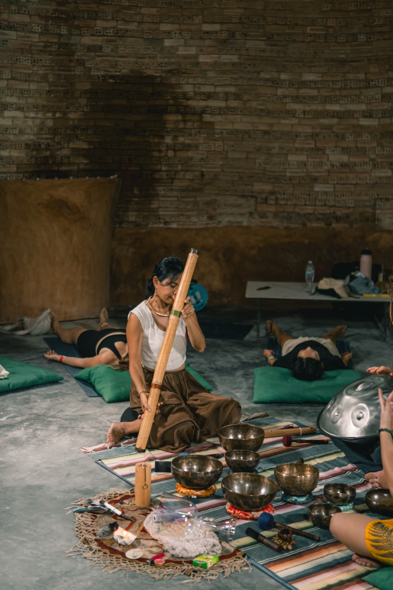 A practitioner kneels holding a tall bamboo didgeridoo beside an arrangement of singing bowls while others rest nearby.