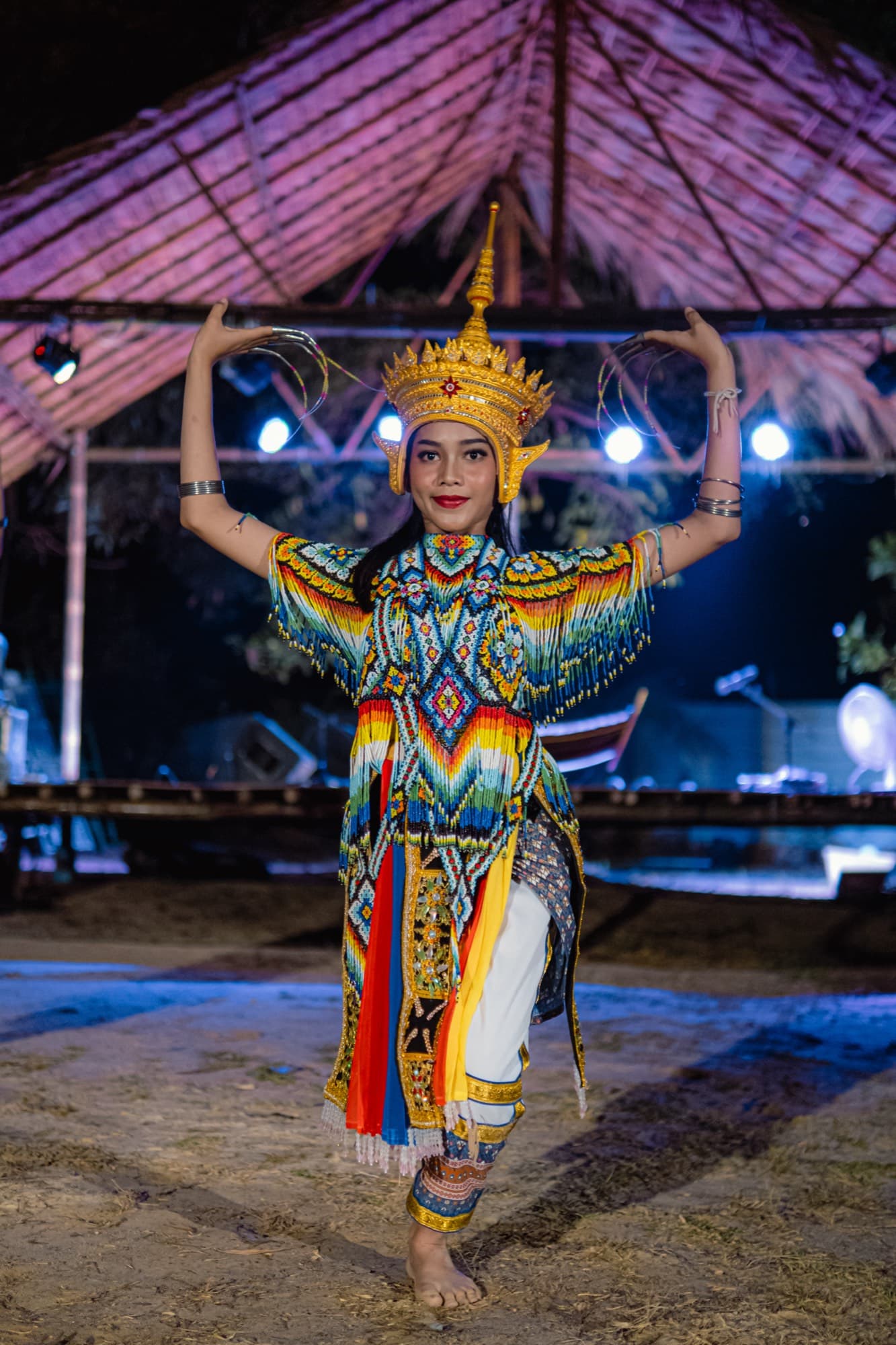 A dancer in a beaded costume and gold crown raises both arms in a classical pose on sand at night.