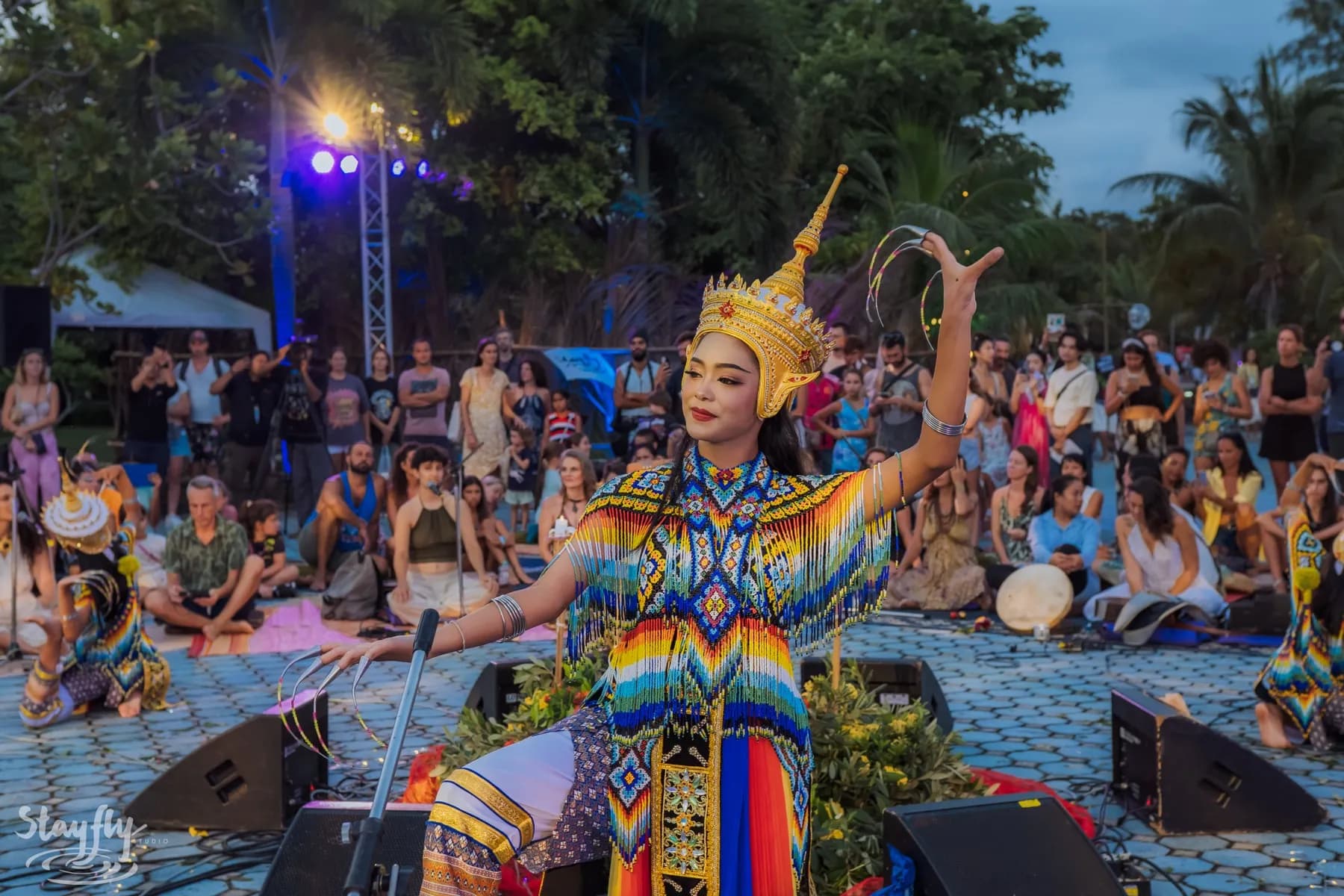 A dancer in a beaded costume and gold headdress performing in the open air, with people watching from the ground.