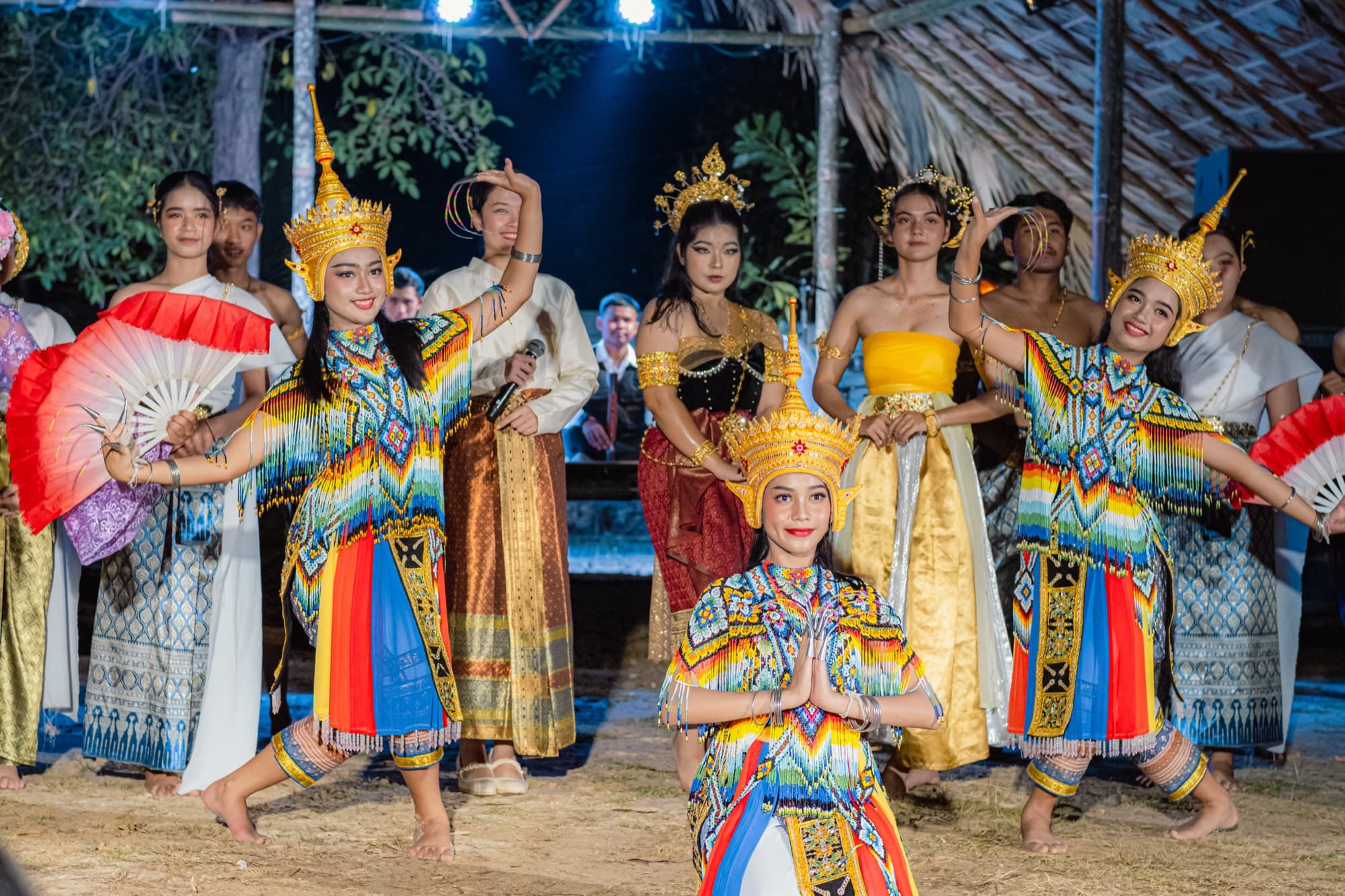 A full troupe of dancers in beaded costume and gold crowns performing together on sand.