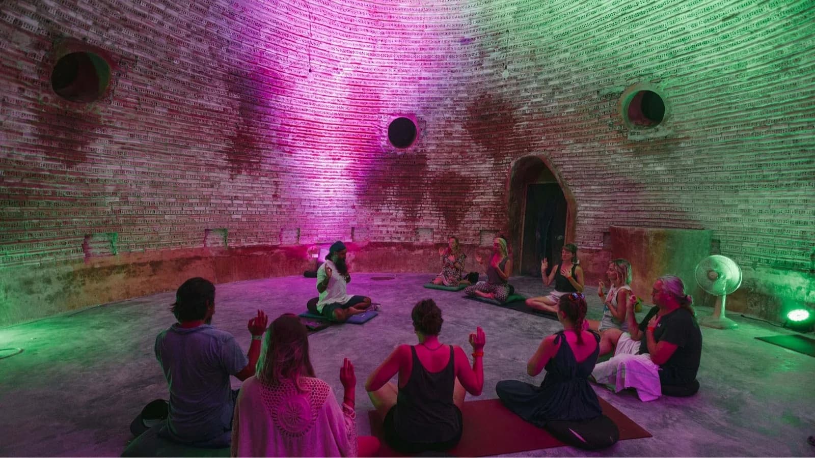 People sitting in a circle on mats inside a round brick room lit in pink and green.