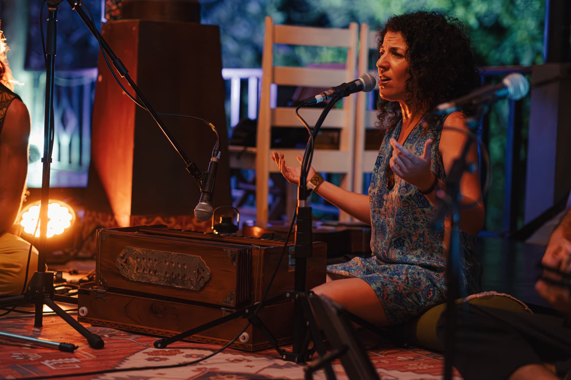 A woman sits cross legged on a patterned rug singing into a microphone beside a wooden harmonium.