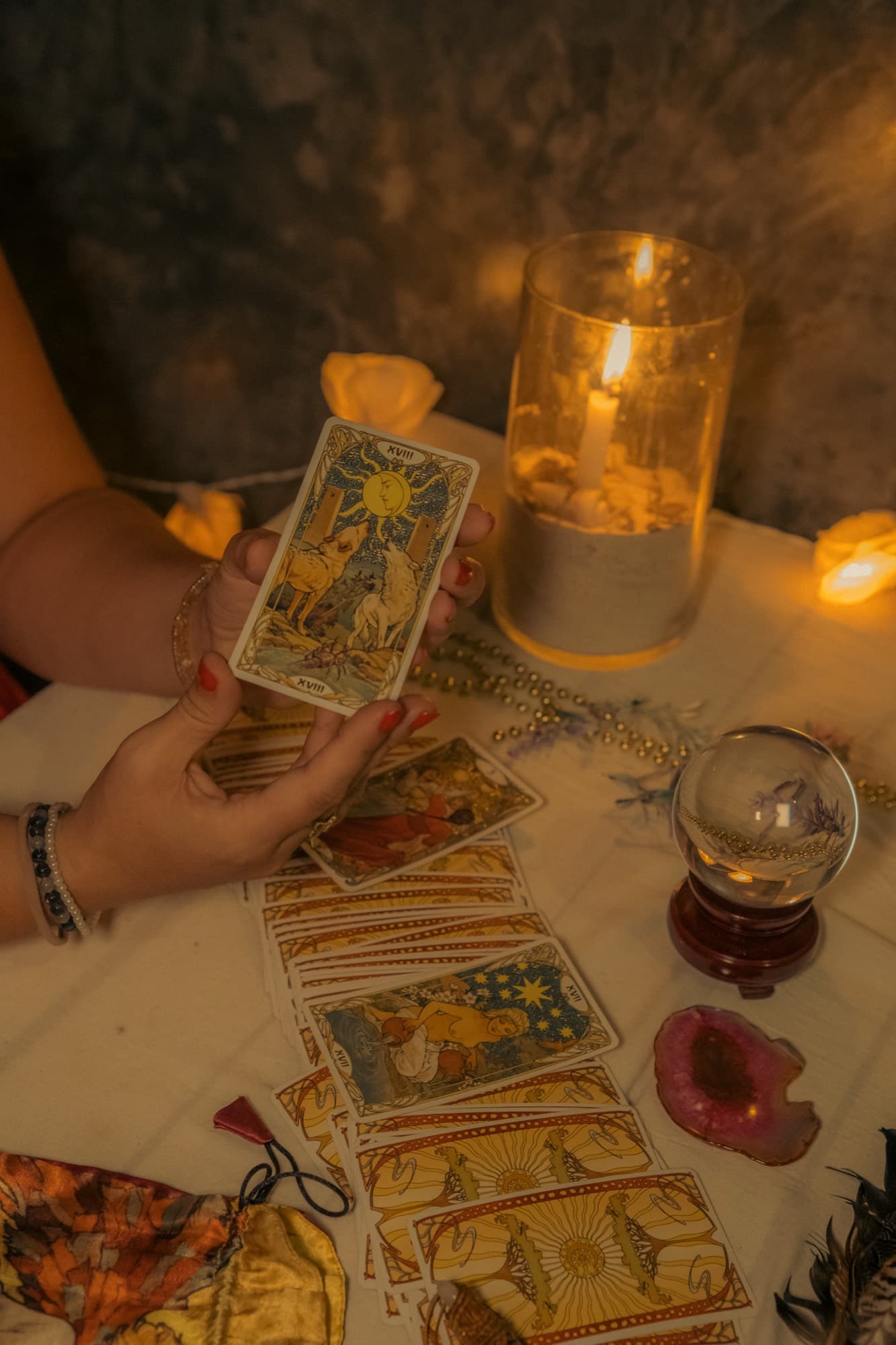 Two hands hold a tarot card above a cloth laid with a spread of cards, a lit candle and a crystal ball on a stand.