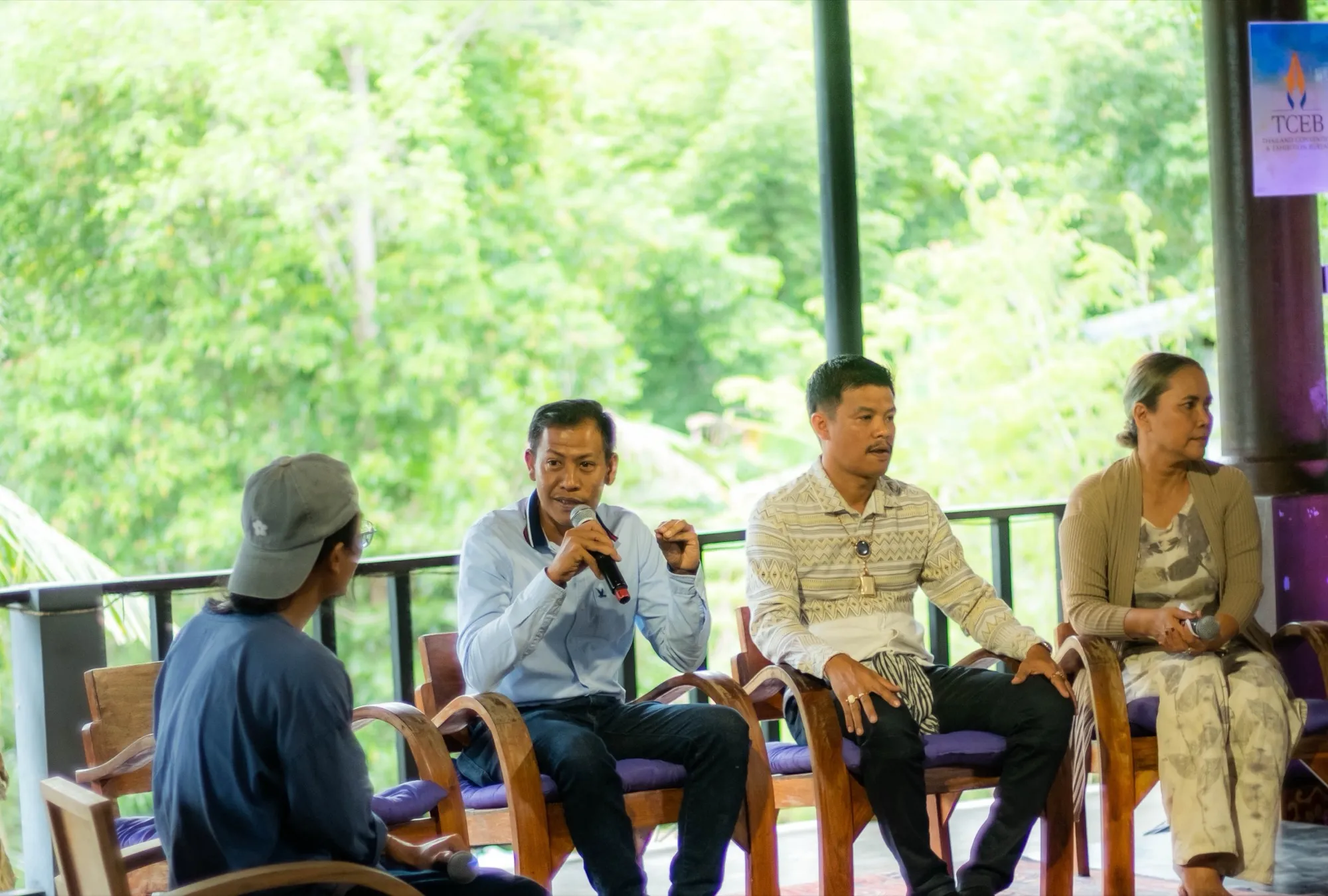 Four people seated in a row of wooden chairs on an open-air deck, one speaking into a handheld microphone, with forest behind them
