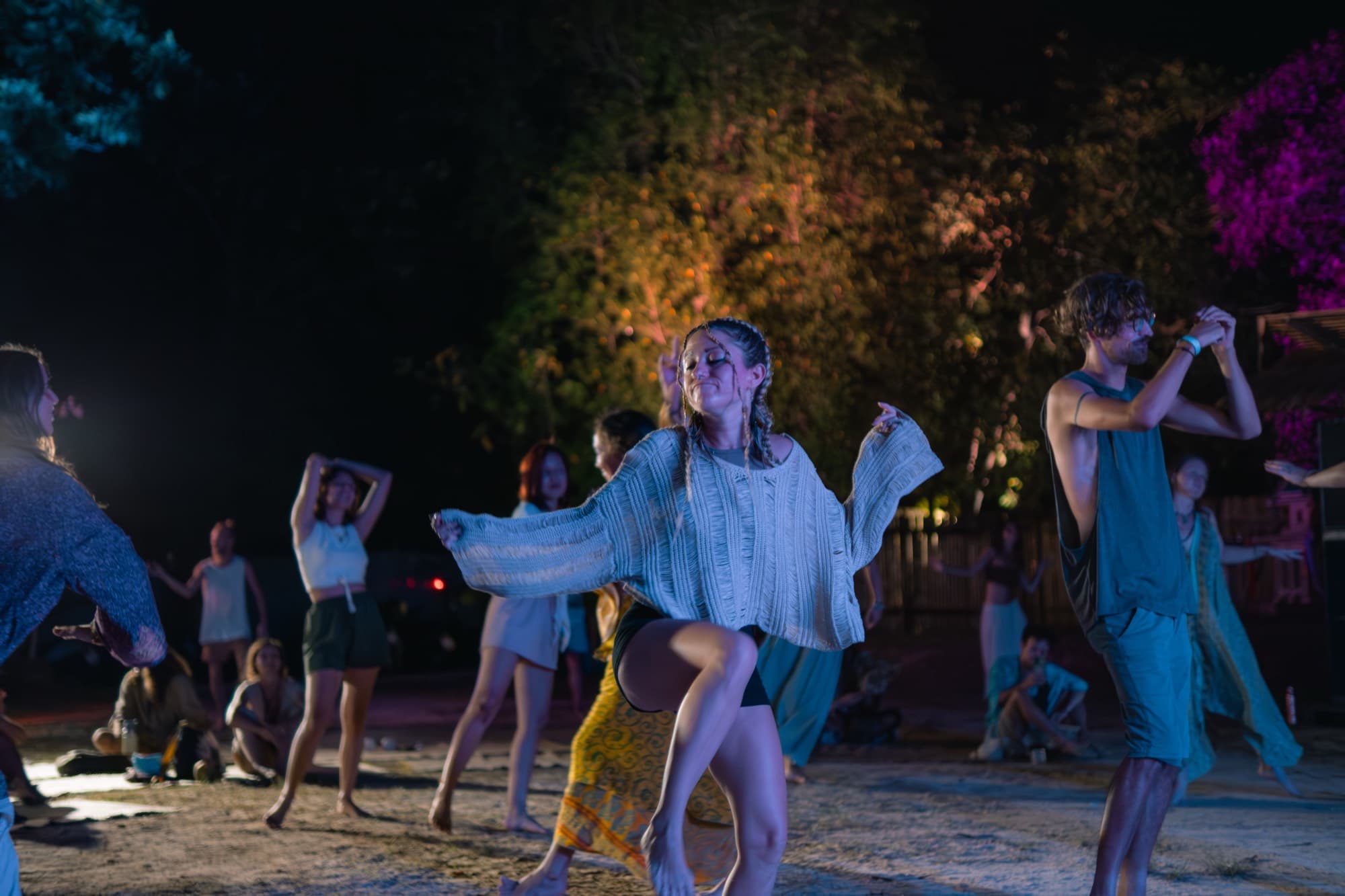 People dancing barefoot on sand at night, the trees behind them lit warm and pink.
