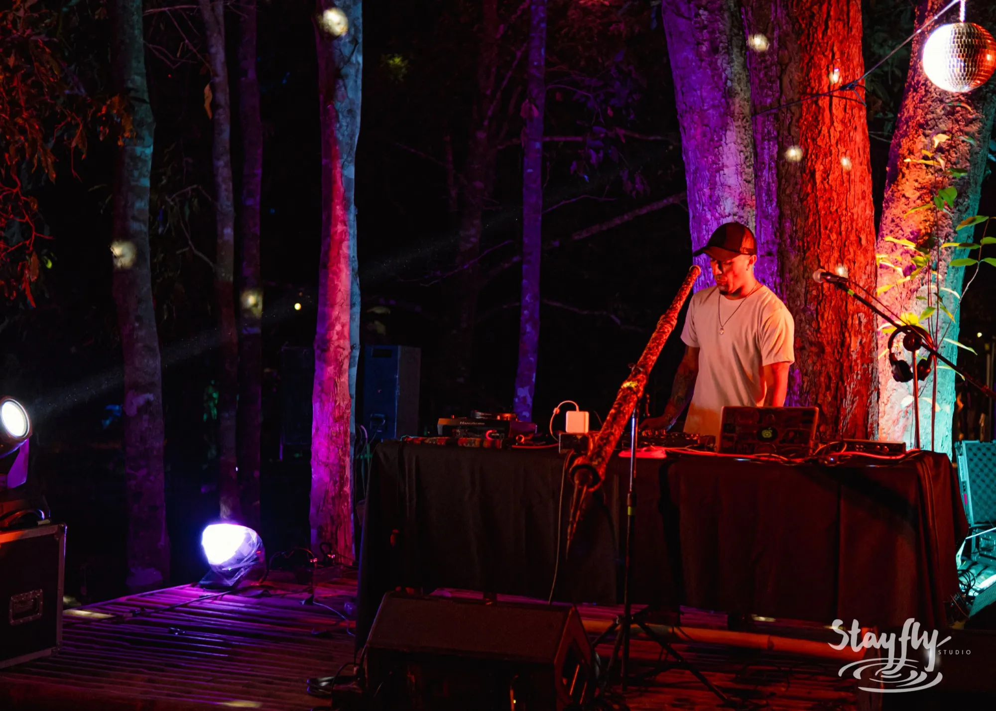 A musician at an outdoor desk of electronic equipment at night, a long wooden wind instrument resting beside him, the trees behind lit red and violet.