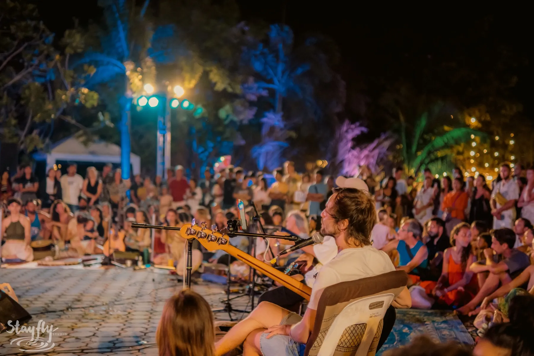A crowd sitting close together outdoors at night, listening to a musician playing a stringed instrument into a microphone.