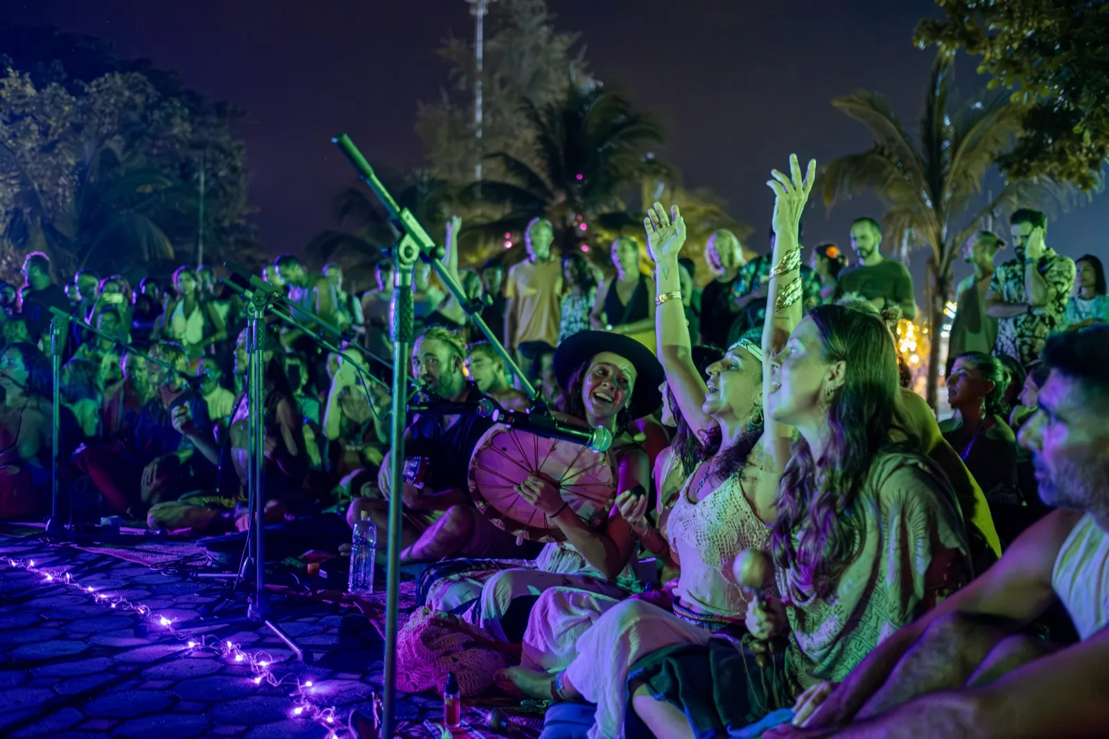 People seated on the ground sing together at night with hands raised, one of them holding a frame drum.