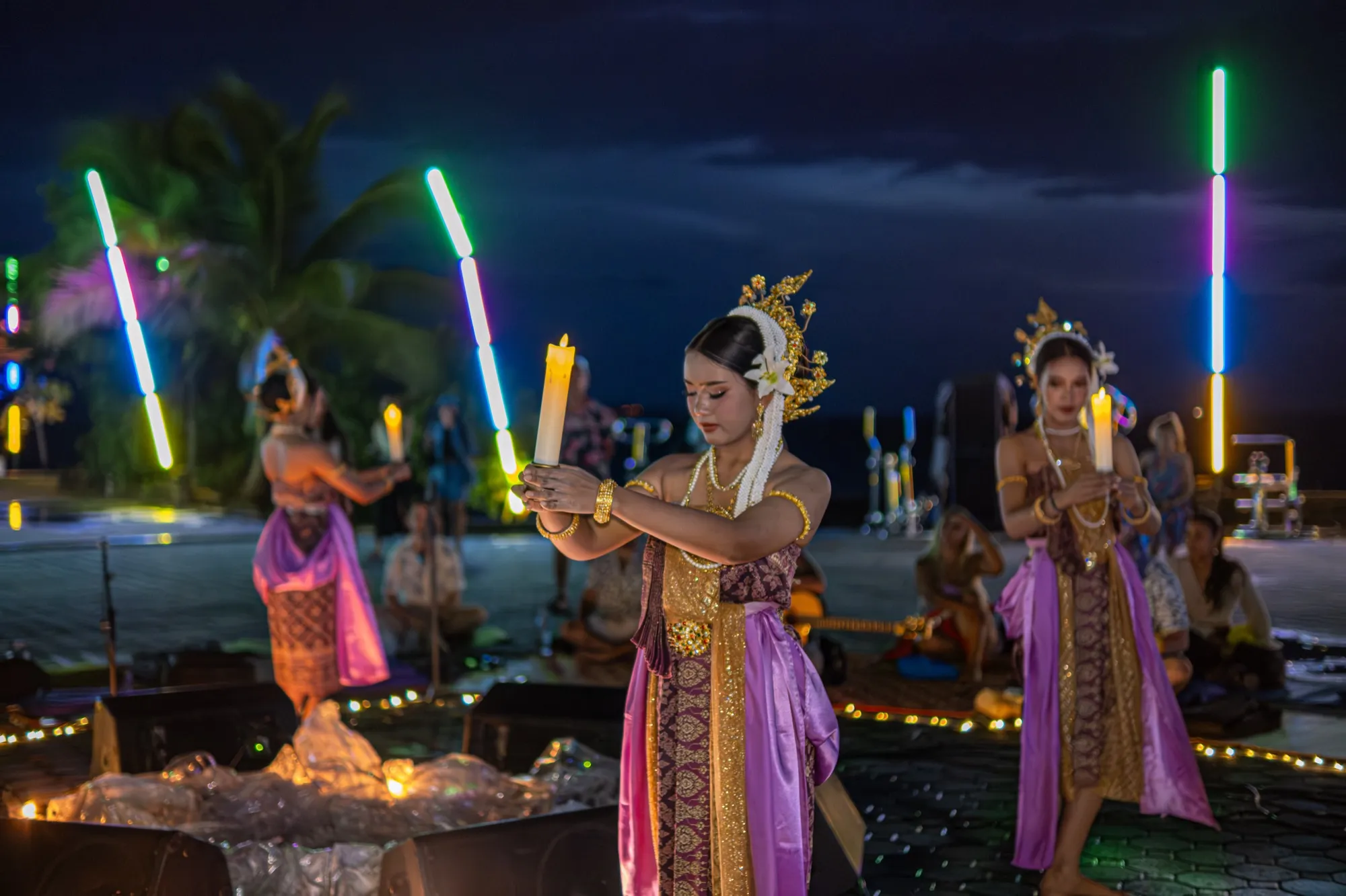 Three performers in gold costume hold candles at dusk beside the sea.