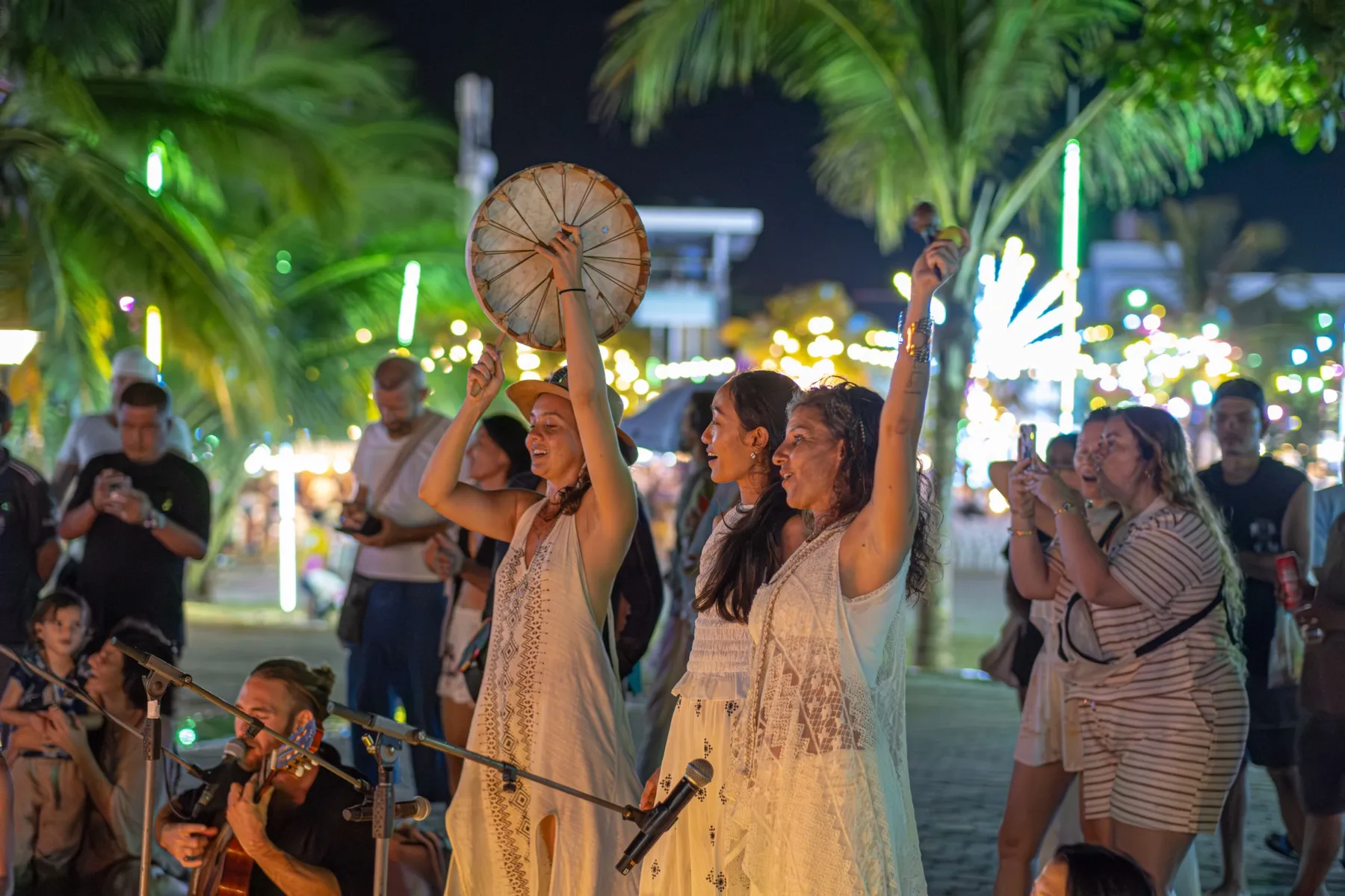 Three women in white raise their arms and lift a frame drum in front of strings of lights at night.