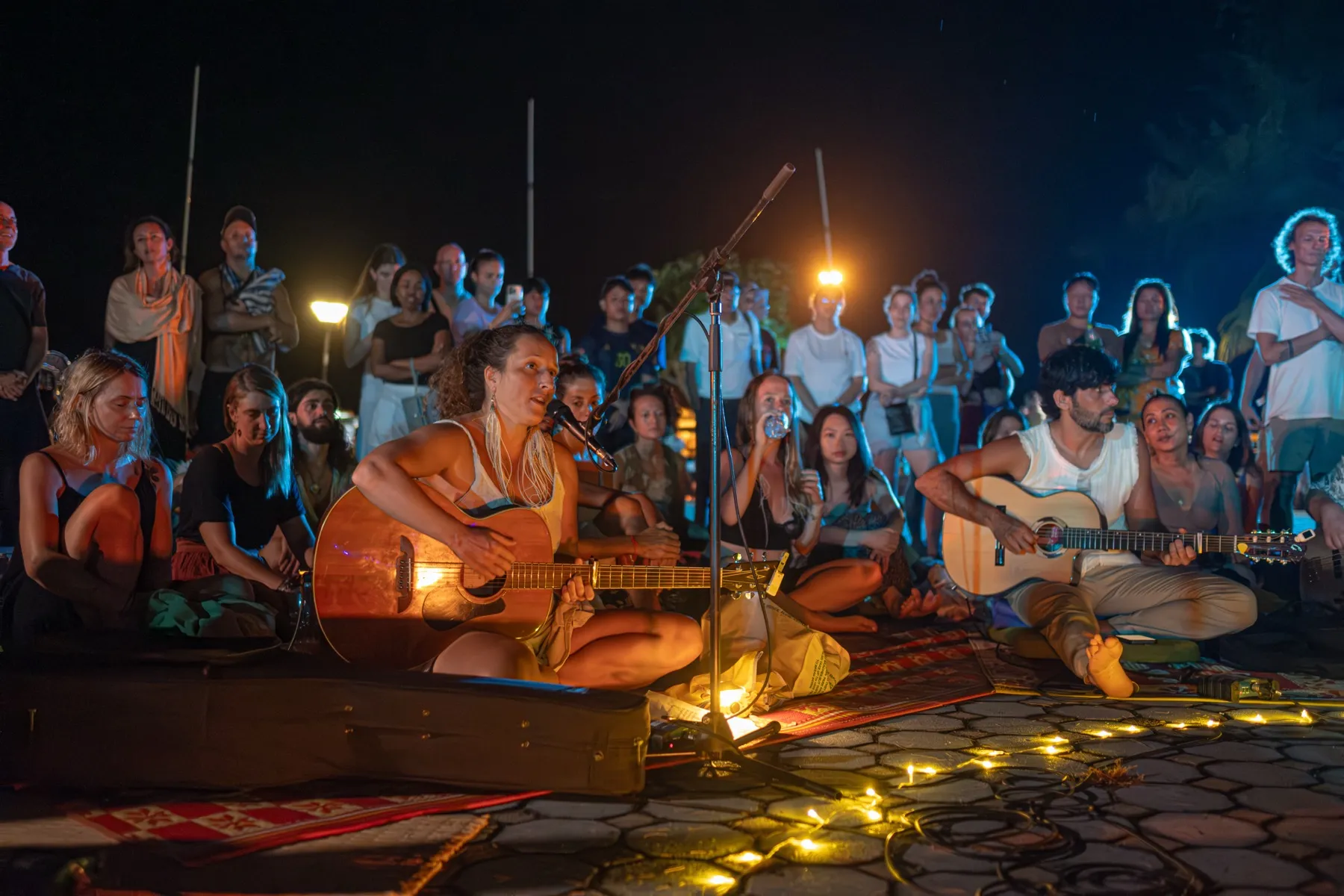 Two guitarists play seated on the ground with the gathering close around them at night.