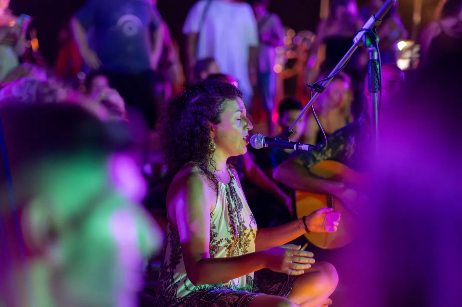 A woman sings into a microphone in profile under coloured light beside a guitarist.