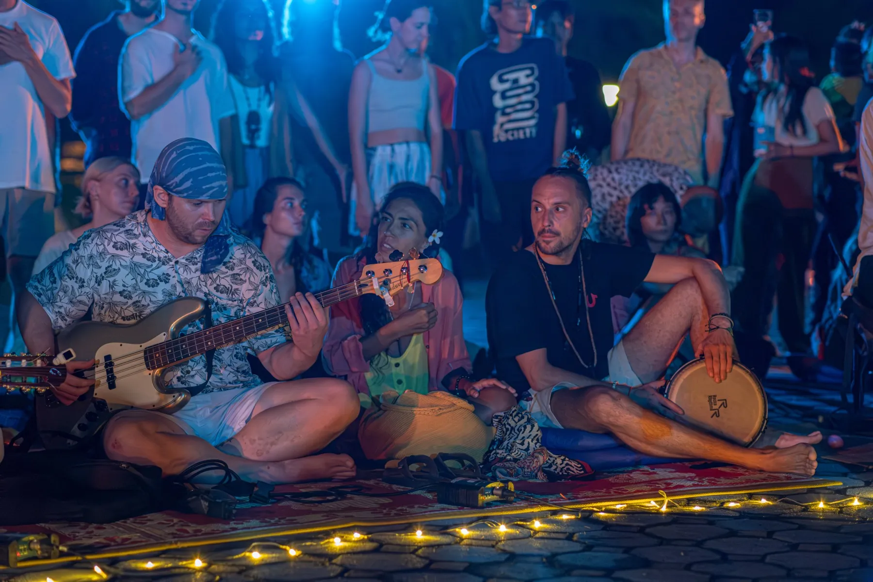 Three musicians play bass, sing and drum while seated together on a rug lit by fairy lights.