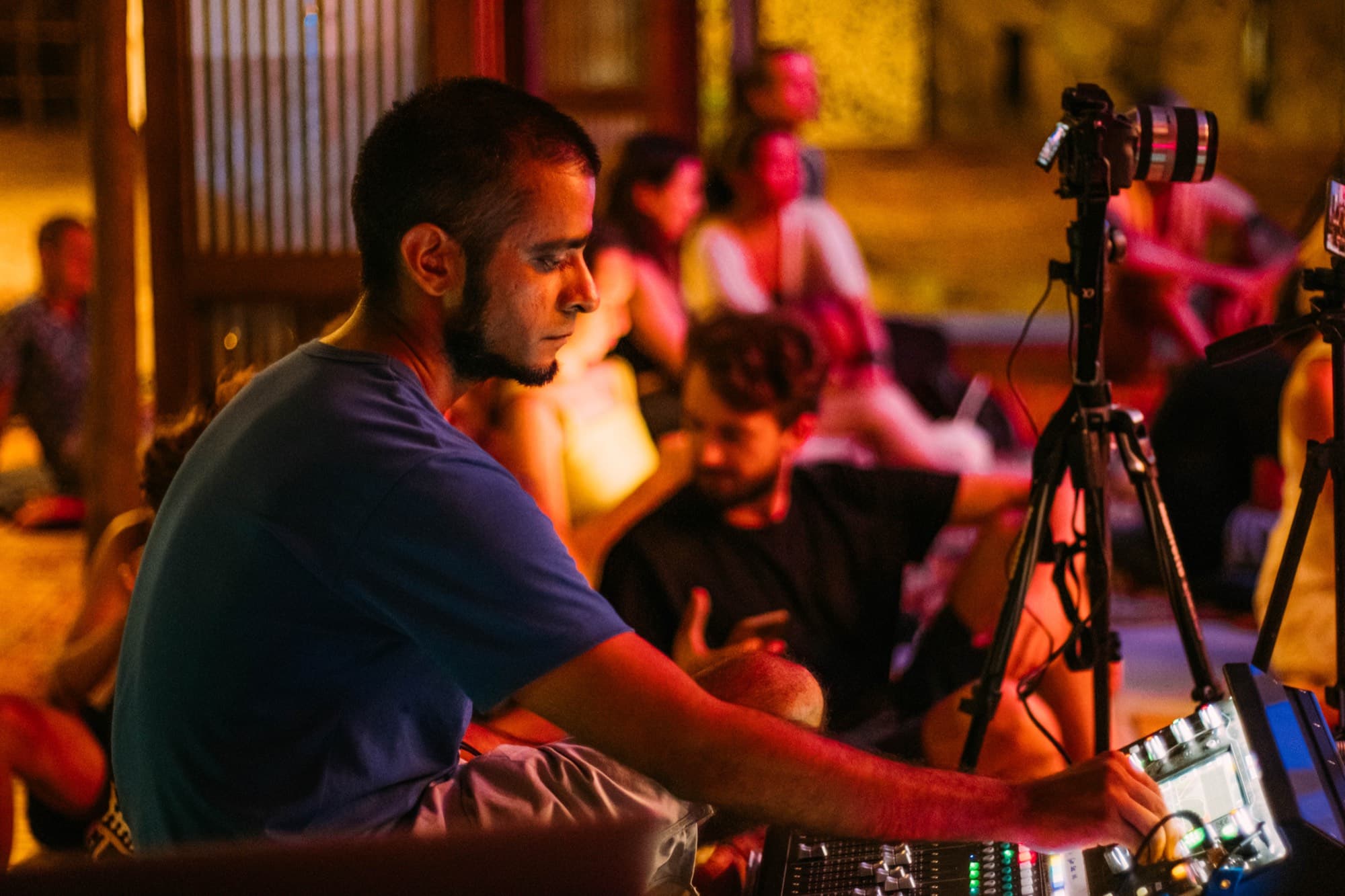 Someone works at the mixing desk while people sit listening behind, a camera recording on a tripod.