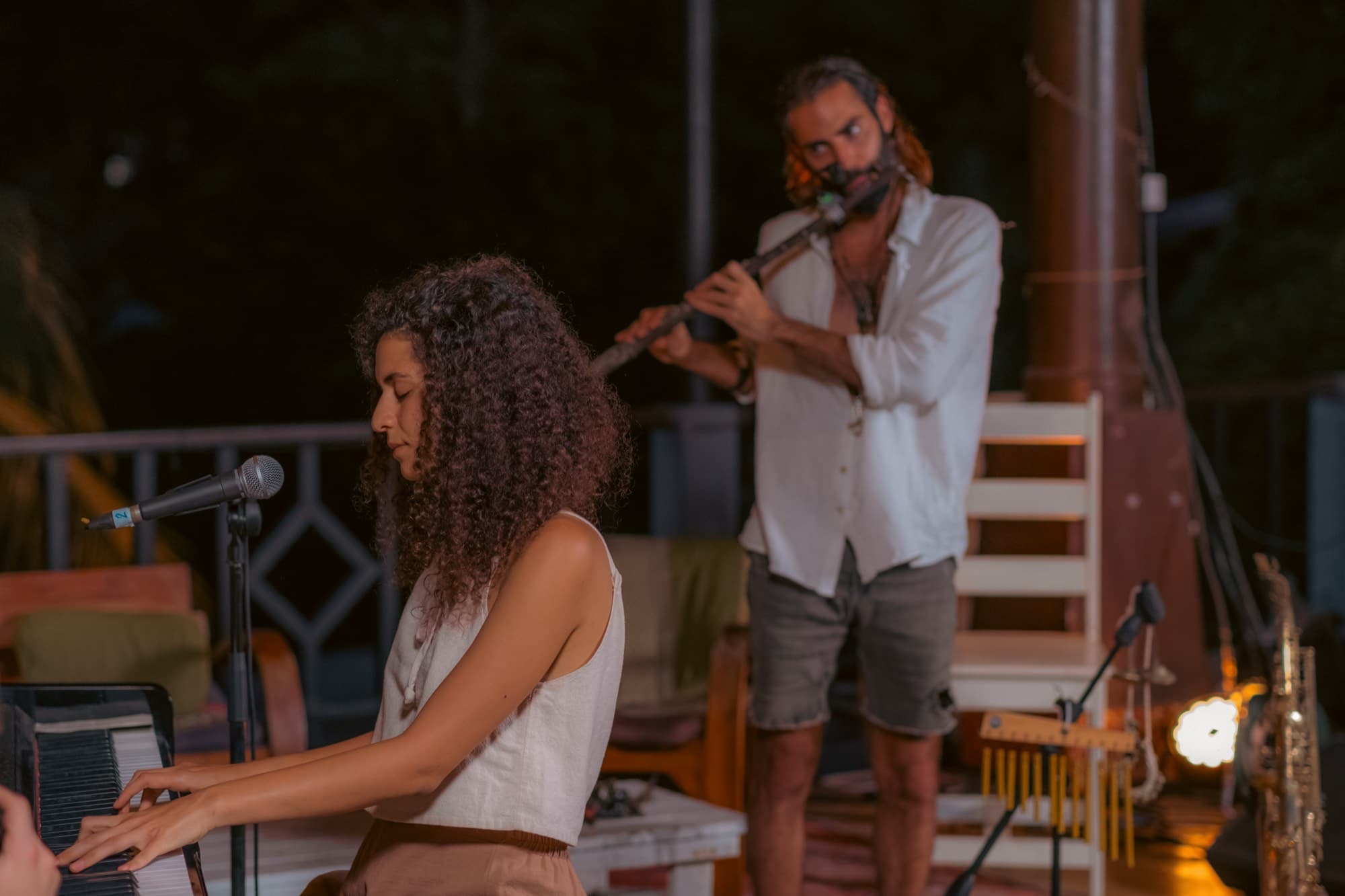 A woman plays a keyboard while a man plays a flute beside her at dusk, a saxophone waiting on its stand.
