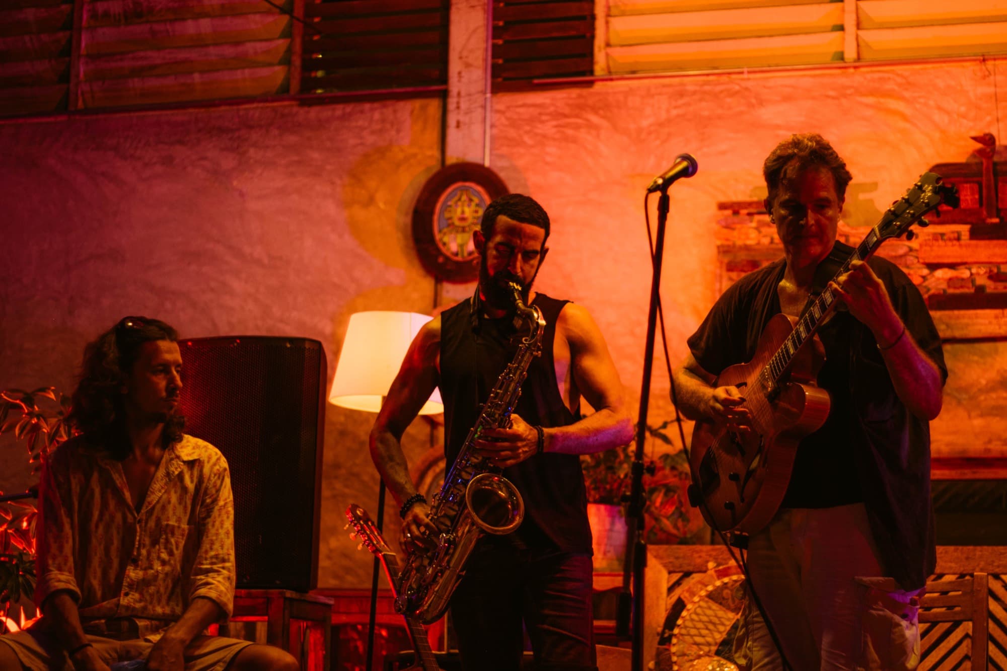 A saxophonist and a guitarist play together in the room under warm light, a third musician listening behind them.