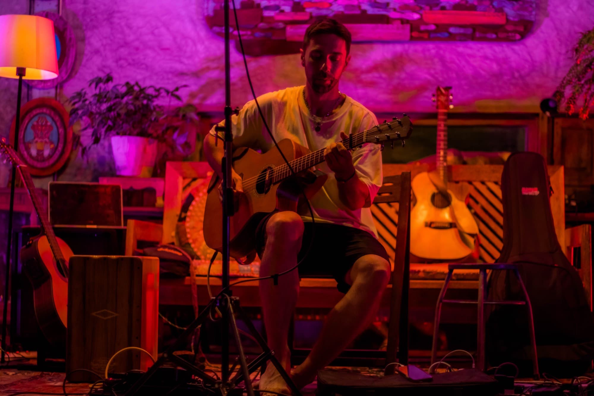 A man sits on a bench playing an acoustic guitar beside a second guitar on a stand and an open case.