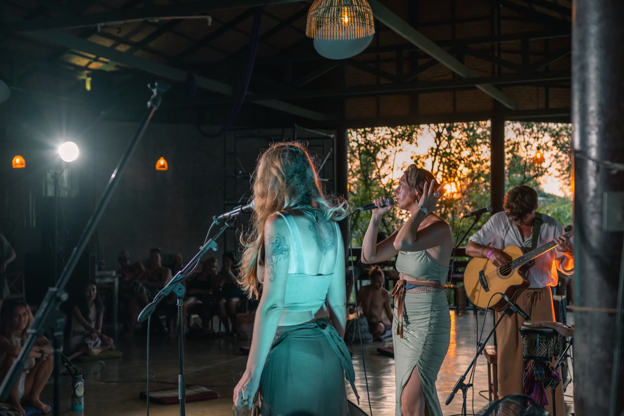 Two women singing into microphones in an open sided pavilion at sunset, a small audience seated on the floor.