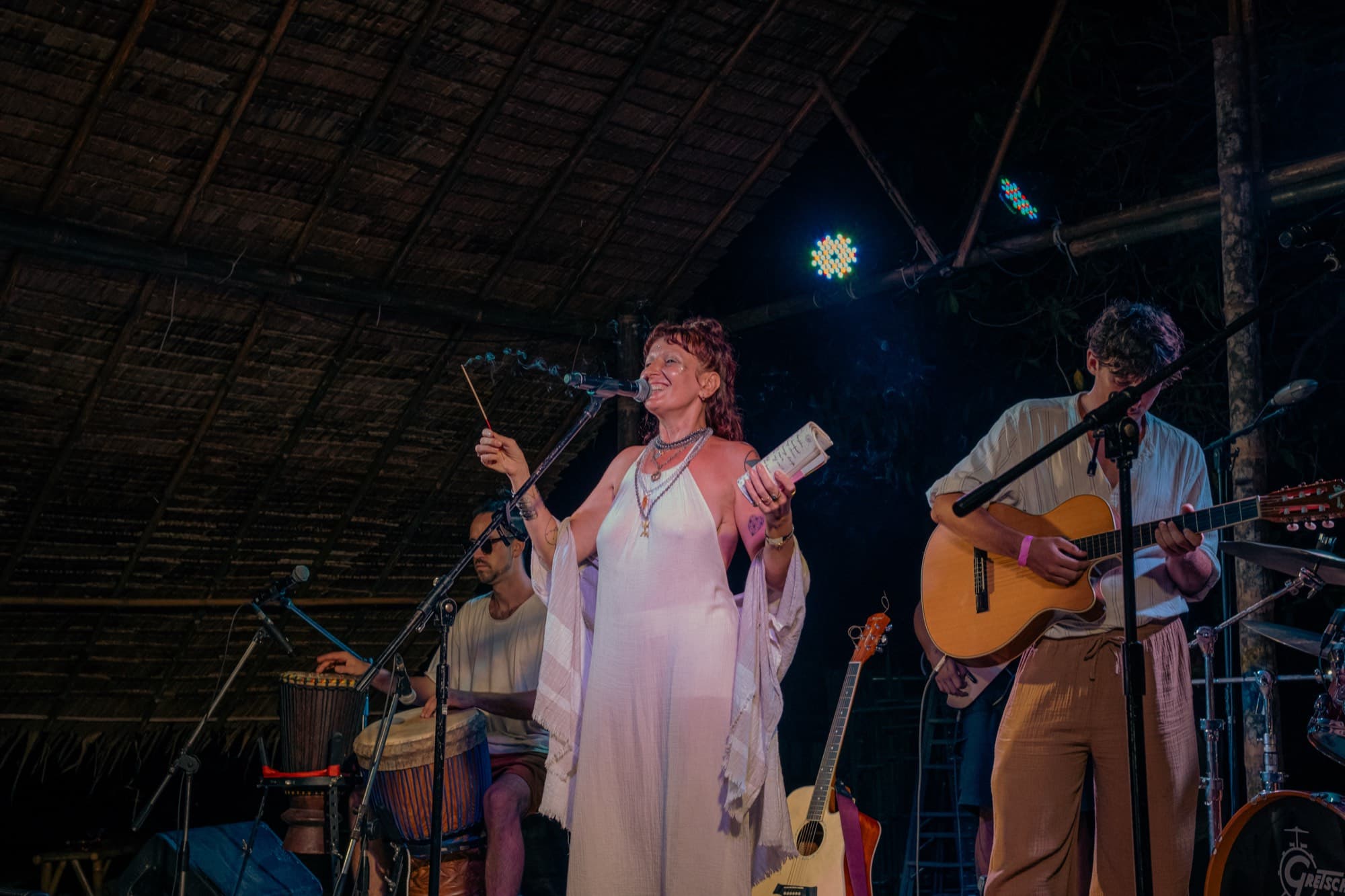 A woman singing into a microphone under a thatched roof at night, a drummer and a guitarist playing beside her.