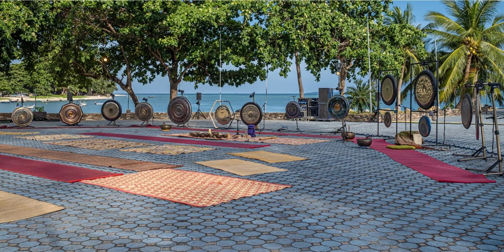 A row of gongs on stands beside woven mats laid out on paving by the sea.