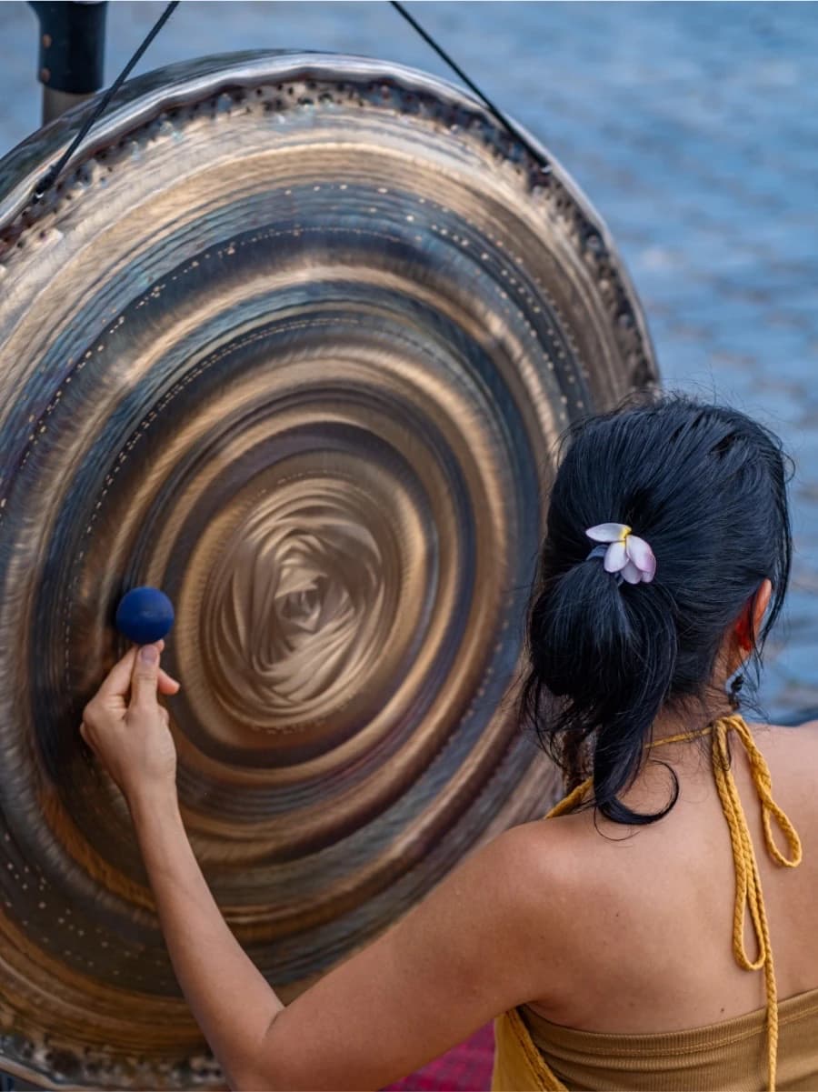 A woman seen from behind lifts a felt mallet to the face of a large gong.