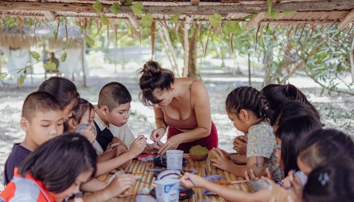 An adult leaning over a long bamboo table to help a group of children with a craft project