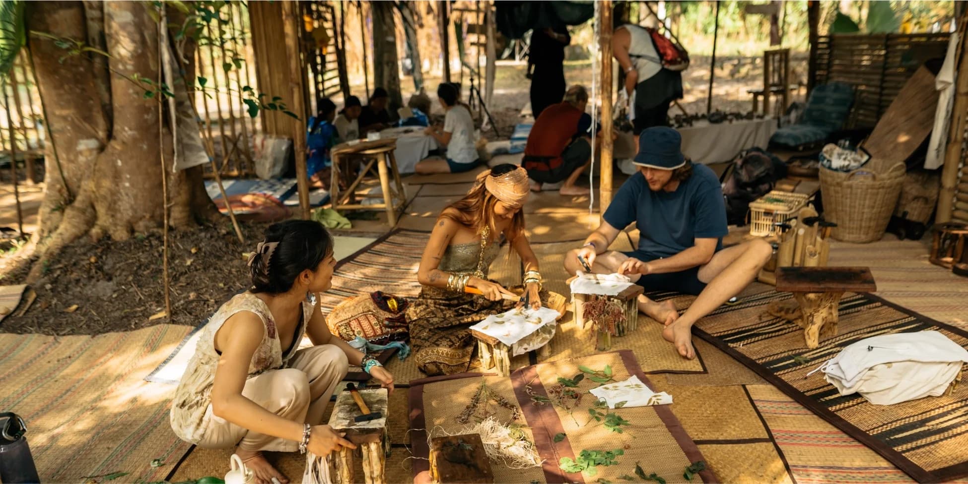 Three people sitting on woven mats in an open bamboo shelter, making things by hand