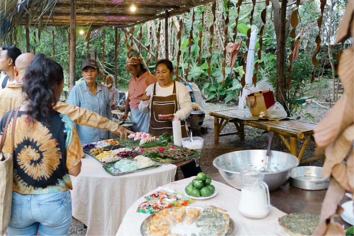 Three people serving homemade dishes from a long table to visitors at an outdoor market