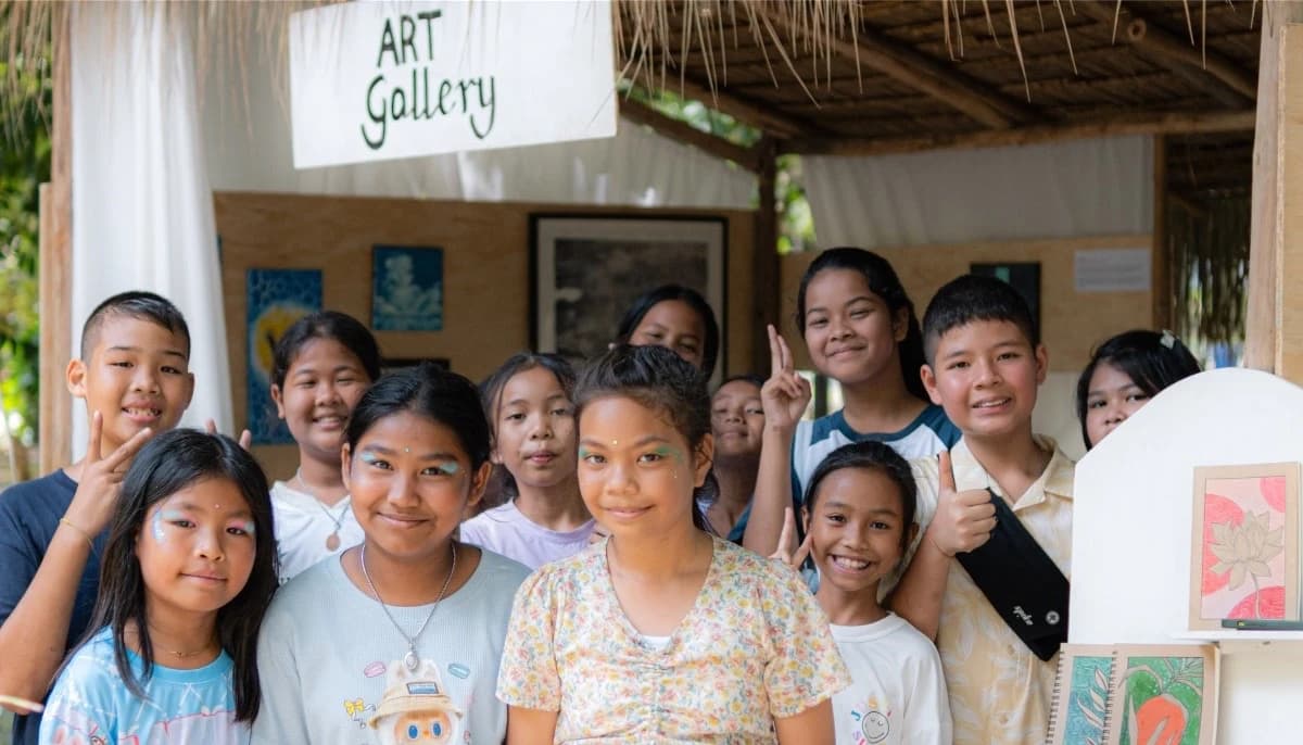 A group of children stand smiling beneath a hand painted Art Gallery sign, with their own drawings displayed behind them.
