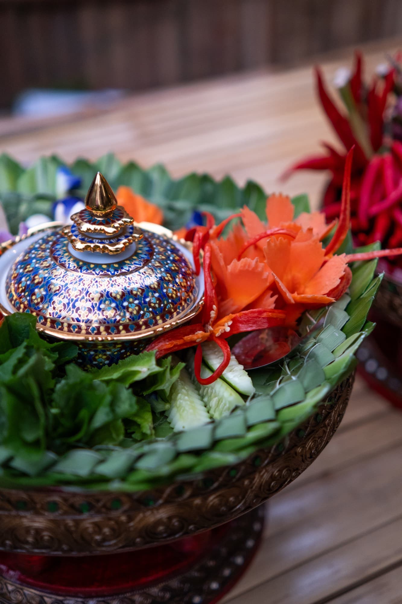 A brass footed tray holding a blue and gold lidded bowl, carved carrot flowers, red chillies, cucumber and lettuce on a woven banana leaf rim.