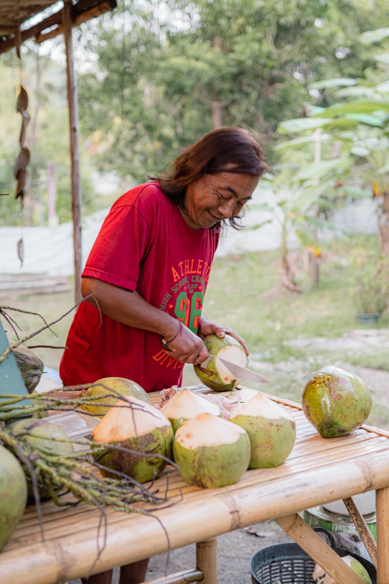 A woman in a red shirt opens a young coconut with a knife at a bamboo table stacked with more coconuts.
