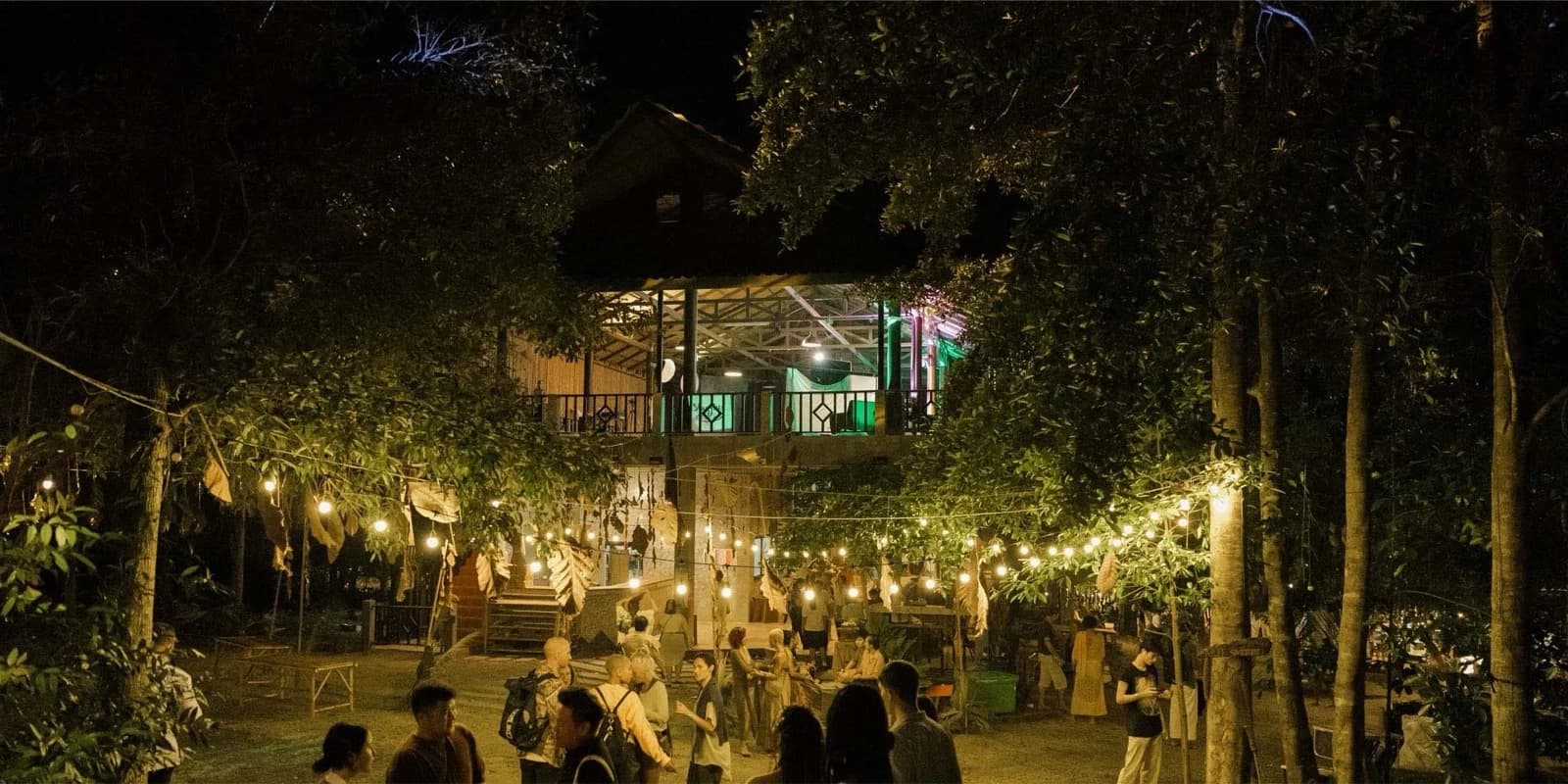 Strings of bulbs lit between trees at night, with a two storey building behind and people walking on the sand.