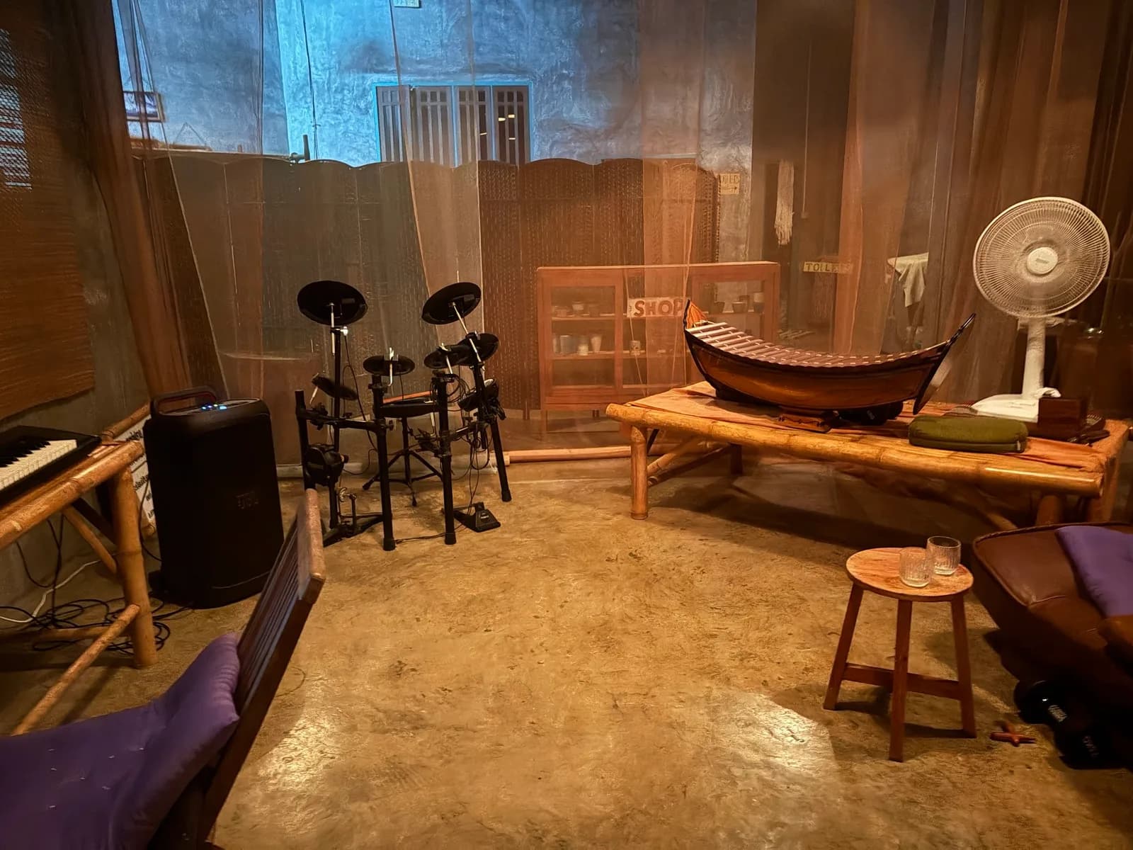 Electronic drum kit, a Thai xylophone on a bamboo table and mesh walls in the open rehearsal room.
