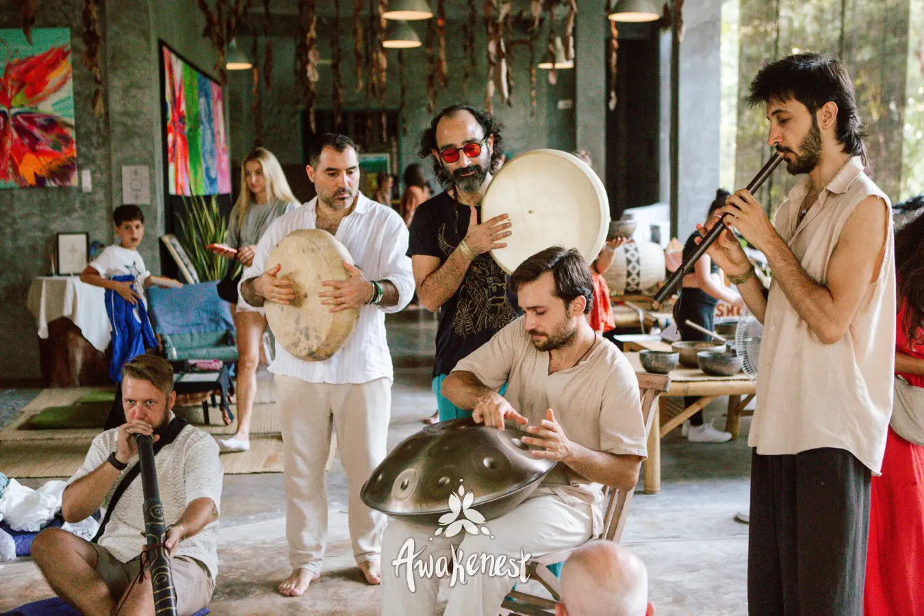 Musicians play frame drums, a handpan, a flute and a didgeridoo together in daylight inside the Music Lounge, paintings on the wall behind them.